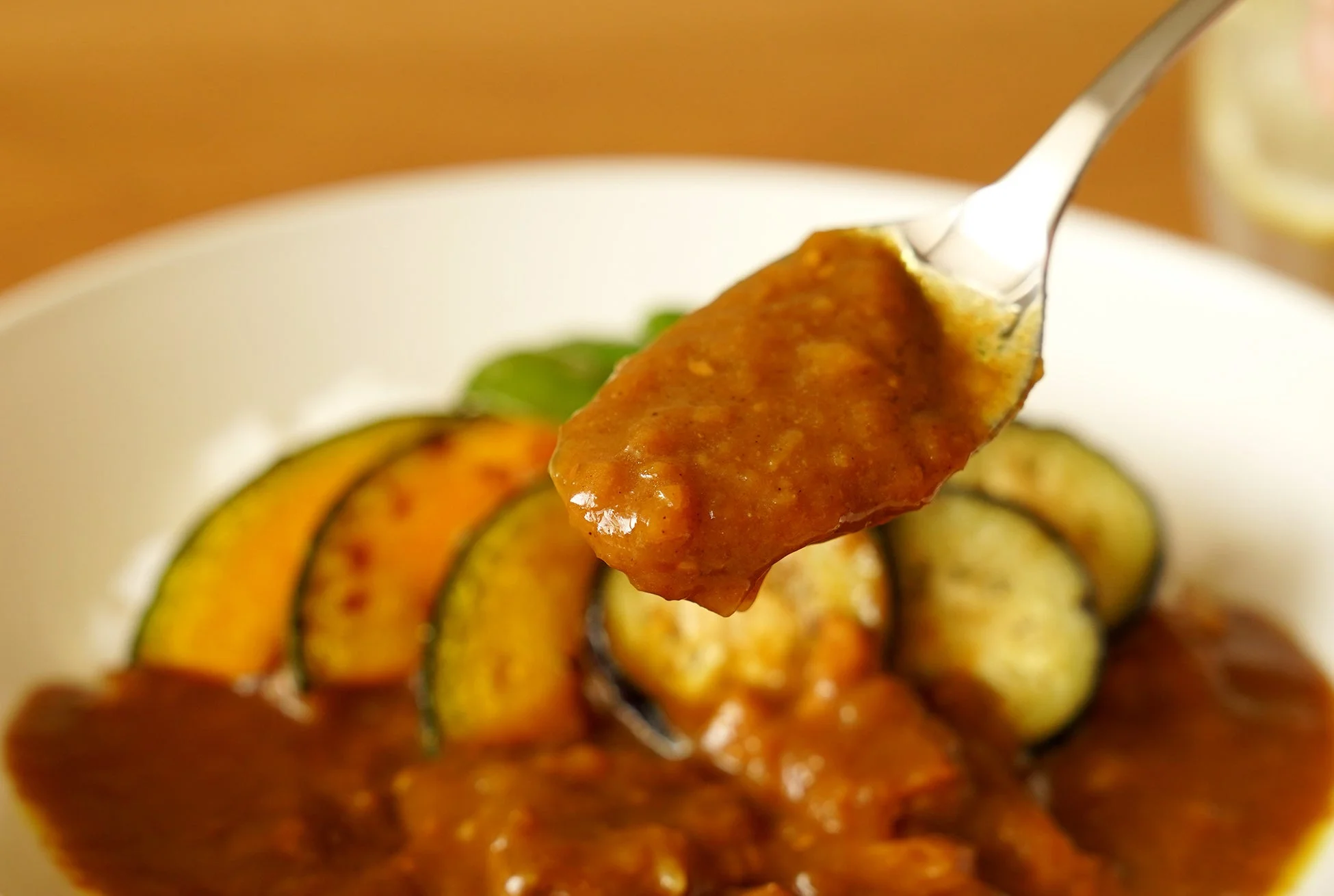スプーンで持ち上げられたとろみのあるカレーがクローズアップで写っています。皿にはご飯と、グリルされたかぼちゃのような彩り豊かな野菜が添えられており、温かい食事が感じられる一枚です。