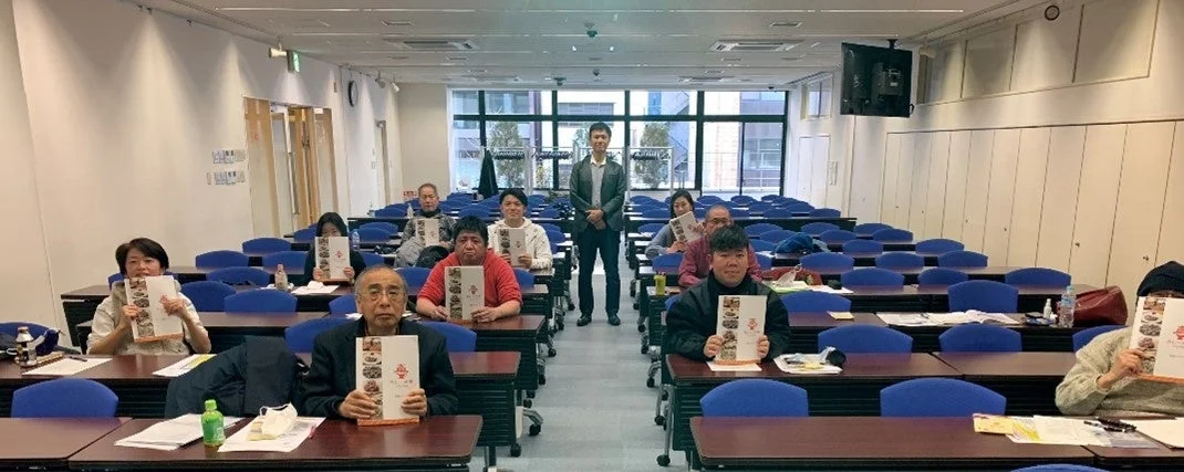 教室のような場所で、複数の参加者が机に座り、「ふるさと納税」と書かれたカタログのような資料を掲げている