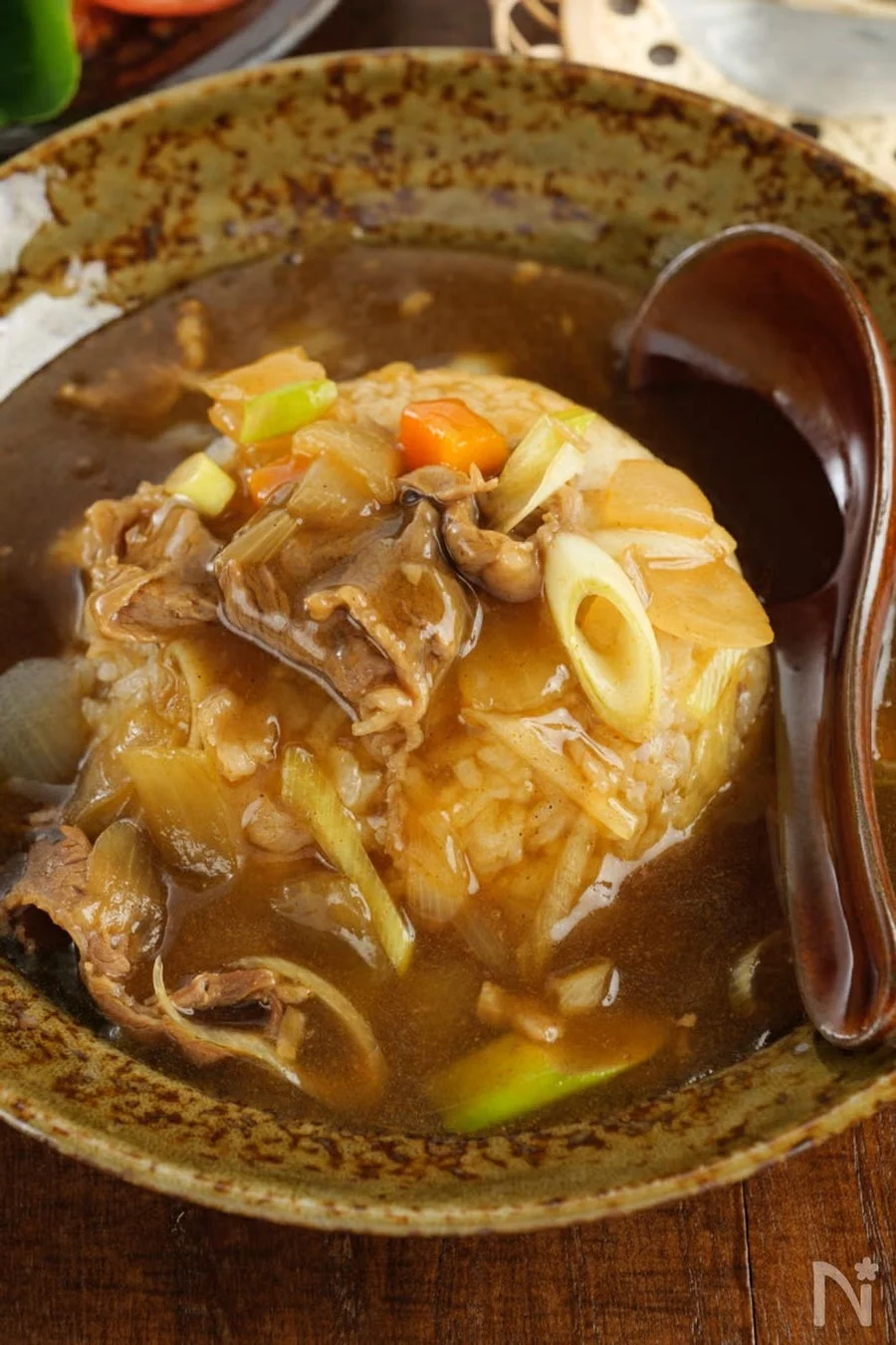 丼鉢に盛られたご飯の上に、牛肉、玉ねぎ、人参、長ネギなどが煮込まれた、とろみのある茶色いあんかけがたっぷりとかけられています。隣には木製の大きなスプーンが添えられており、温かい食欲をそそる一品です。
