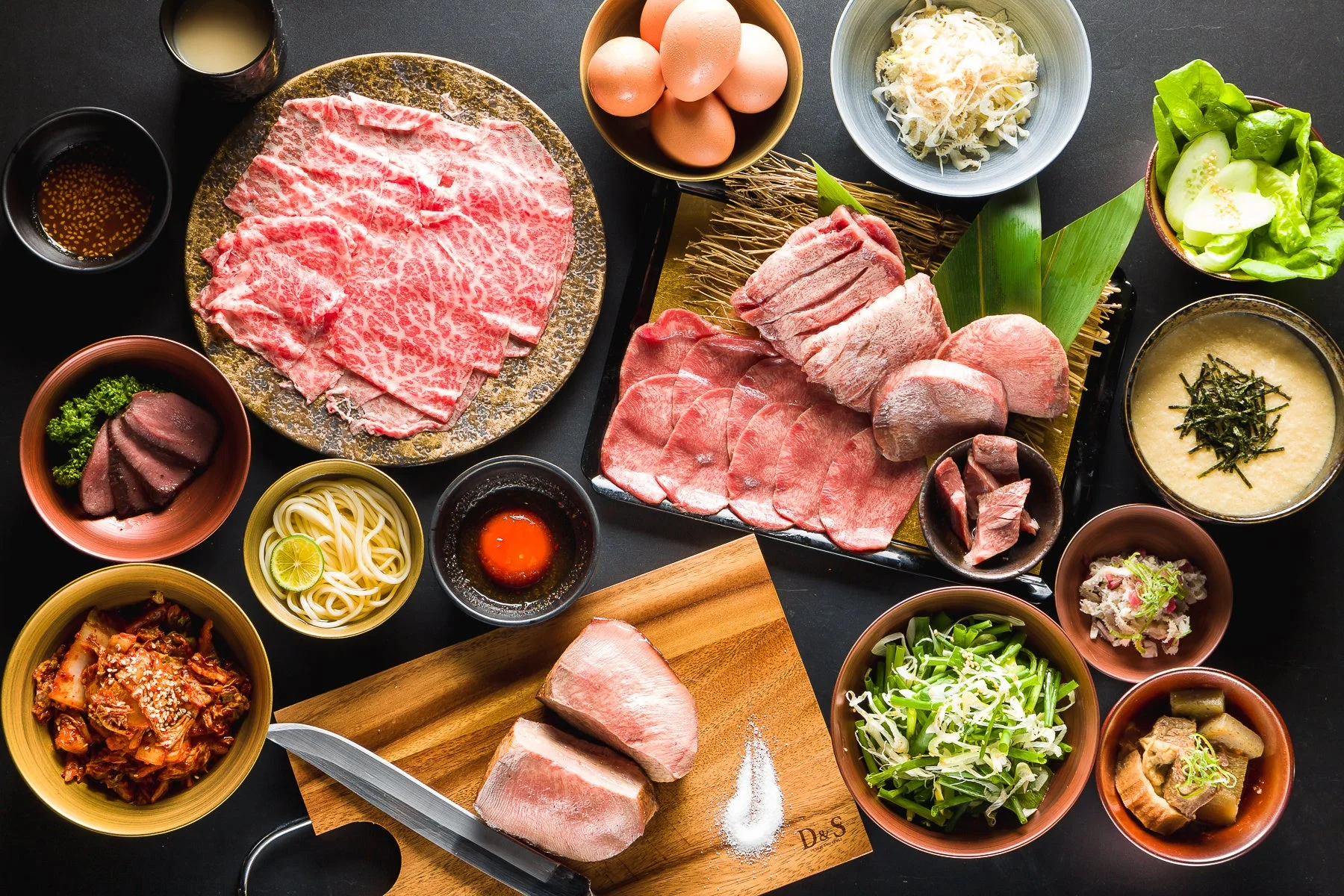 豪華な牛肉と牛タンの盛り合わせを中心に、キムチ、卵、麺類、様々な副菜が並べられた食卓の俯瞰写真です。焼肉やすき焼きを思わせる、彩り豊かで食欲をそそる食事の準備風景が写されています。