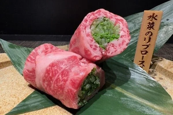 霜降りの美しい牛肉（リブロース）で水菜を巻いた料理が、緑の葉の上に盛り付けられています。