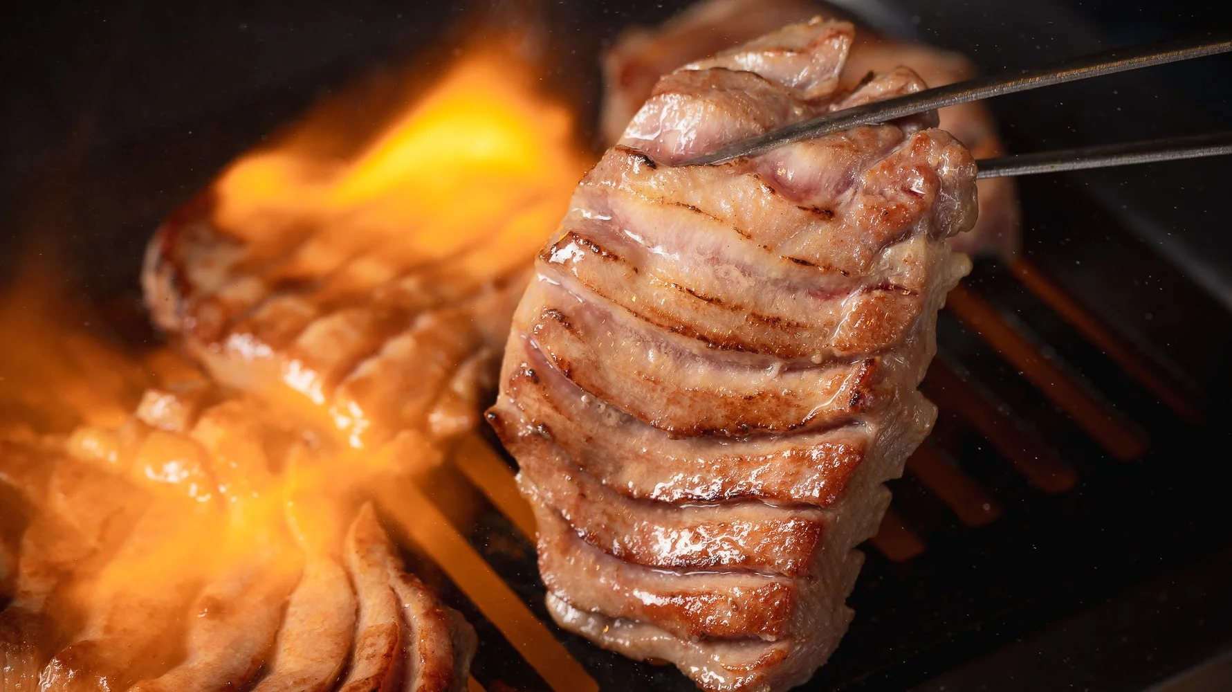 炎が上がる焼き網で、厚切りの牛肉が香ばしく焼かれている様子を捉えたクローズアップ写真。食欲をそそる焼き色が特徴的です。