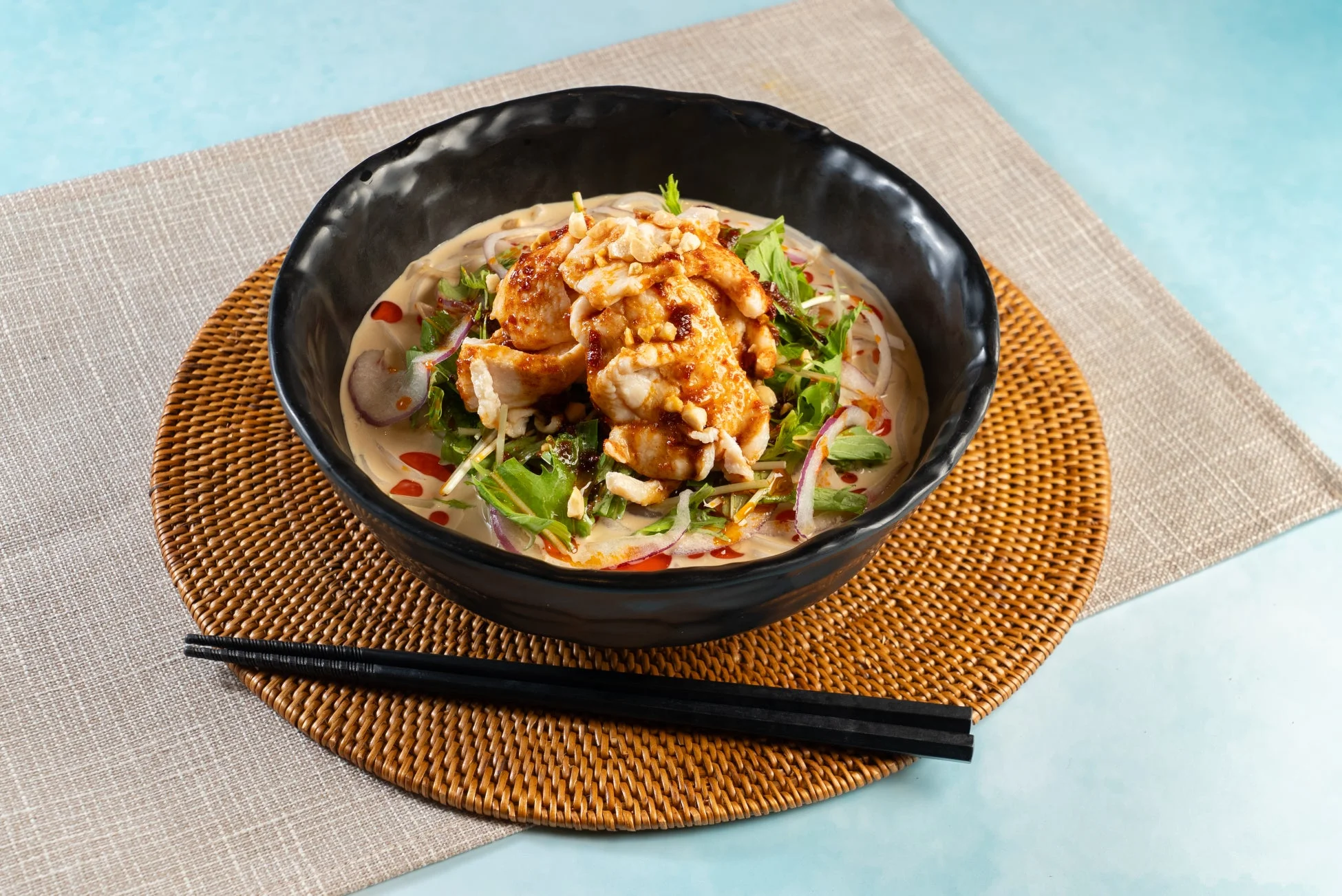 クリーミーなスープに鶏肉、ピーナッツ、ハーブ、赤玉ねぎが盛り付けられたアジアンテイストの麺料理。黒い器に盛られ、籐製のランチョンマットと箸が添えられています。