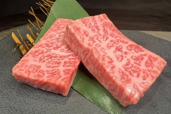 美しい霜降りの入った厚切りの和牛の生肉が2枚、緑の葉を添えた黒い皿に盛り付けられています。