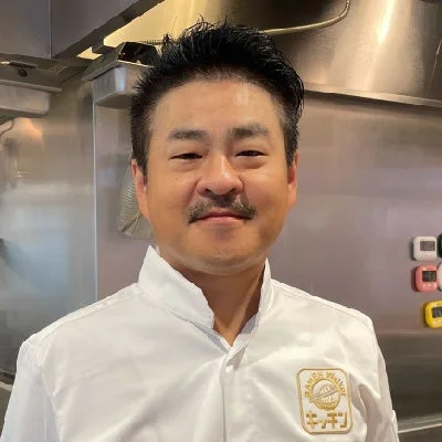 ラーメンWalkerキッチンの厨房で白いシェフ服を着て笑顔の小宮一哲氏