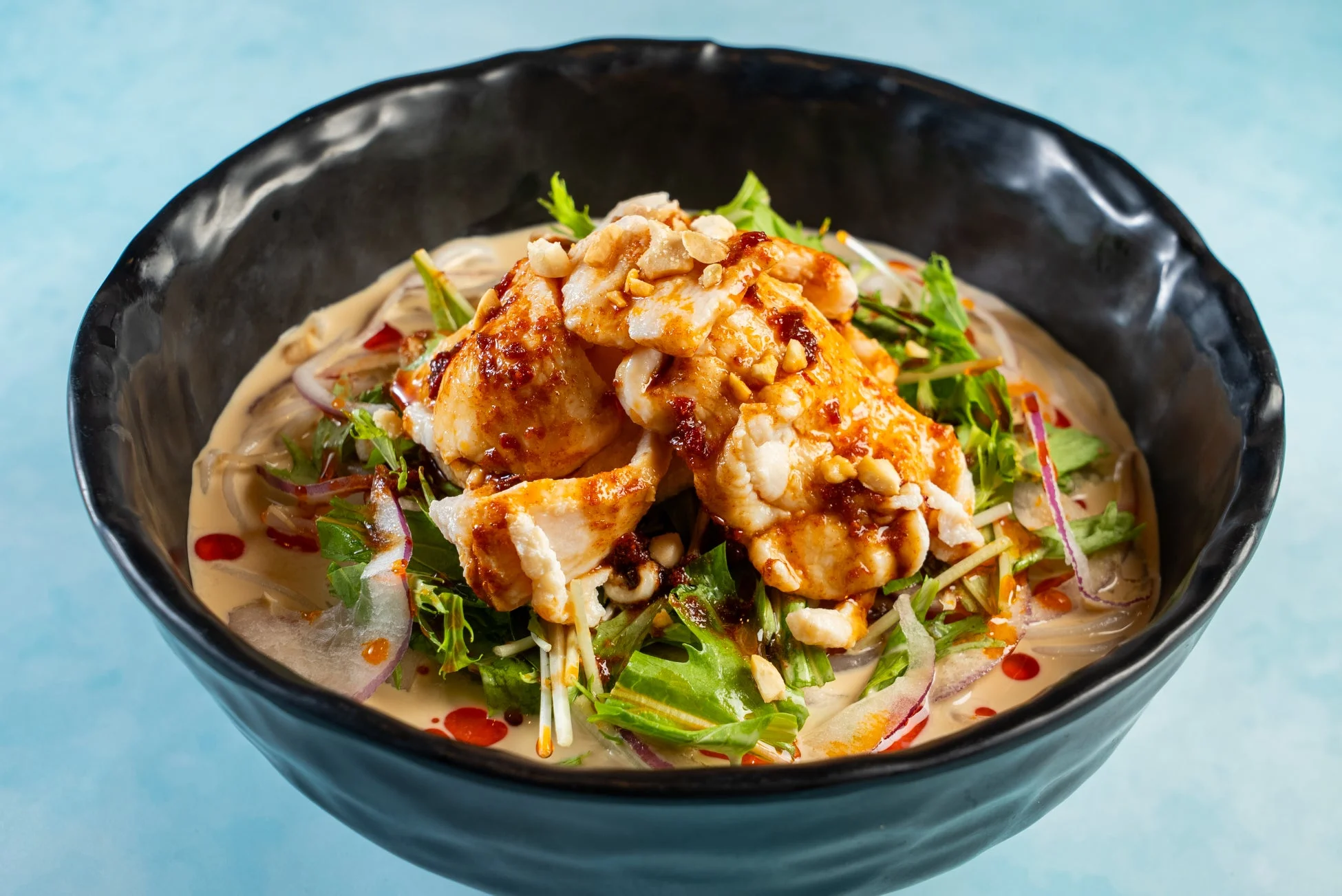 黒い器に盛られた、魚介か鶏肉のピリ辛あんかけ麺料理です。ピーナッツと新鮮な野菜がトッピングされ、クリーミーなスープが特徴的なアジア風のヘルシーな一品です。