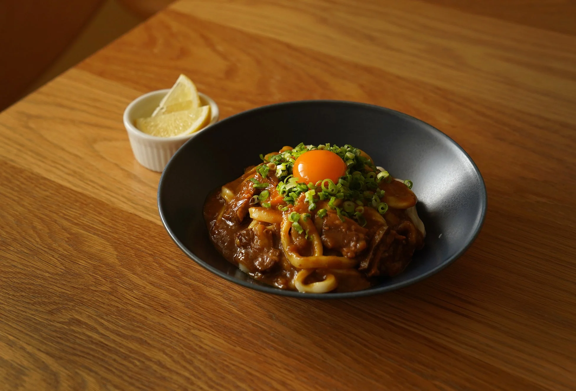 木製のテーブルに置かれた黒い器に入ったカレーうどんです。太い麺に濃厚なカレールーが絡み、中央には鮮やかな卵黄と刻みネギがトッピングされています。横にはレモンが添えられており、食欲をそそる一品です。