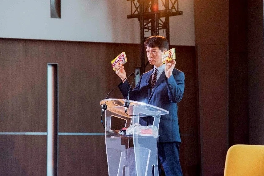 スーツを着た男性が演台の前に立ち、両手に小さな箱を持って聴衆に何かを提示している