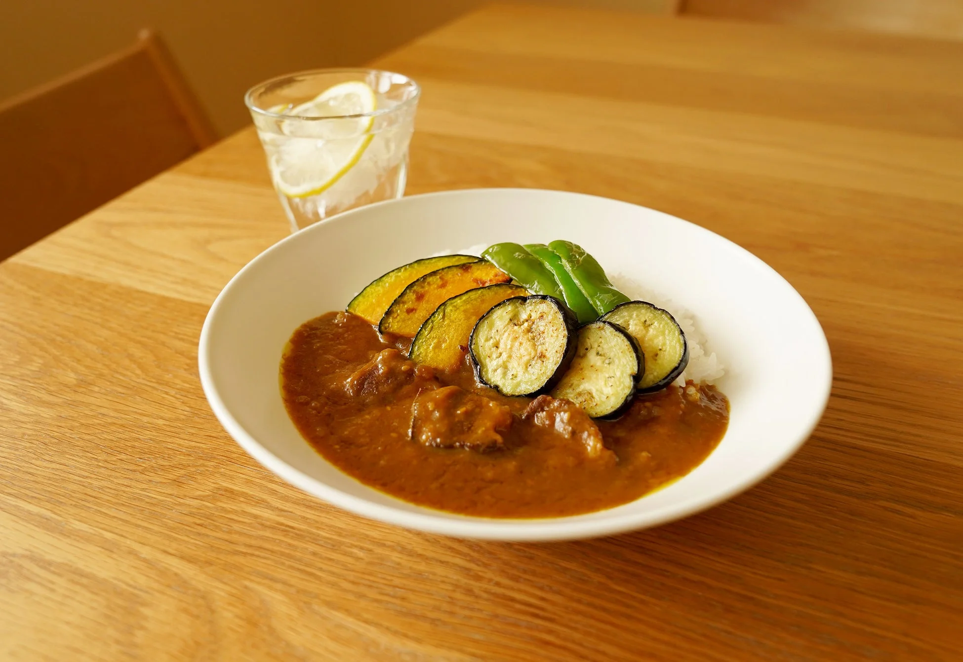 木製のテーブルに置かれた白い皿に盛り付けられた野菜カレーです。ご飯の上に茄子、かぼちゃ、ピーマンが添えられ、その横には牛肉入りのルーがかけられています。奥にはレモンが入ったグラスが置かれており、温かい家庭的な食卓の様子が伺えます。