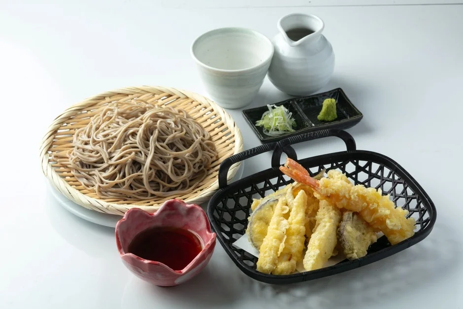 白い背景に置かれた和食のセットで、竹ざるに盛られたそばと、海老や野菜の天ぷらが黒い籠に入っています。花形の器に入ったつゆ、薬味のわさびとネギ、そしてそば湯と思われる白い器と徳利も添えられています。