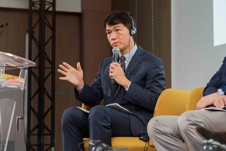 スーツを着たアジア系の男性がヘッドホンを装着し、マイクを持って話している