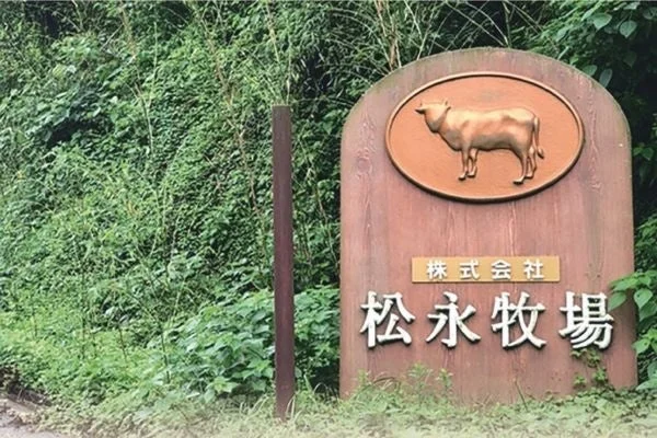 緑豊かな背景の中に立てられた木製の看板が写っています。看板には牛のエンブレムと「株式会社 松永牧場」という文字が書かれています。