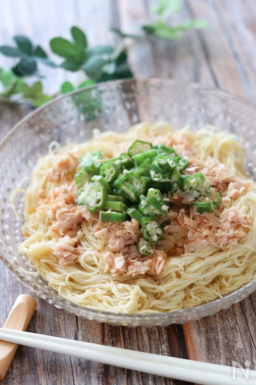 ガラスの器に盛られたそうめんです。細い麺の上に、緑色の輪切りオクラとツナフレークがたっぷりトッピングされており、夏らしいさっぱりとした一品に見えます。木製のテーブルと箸が添えられ、和の食卓を演出しています。