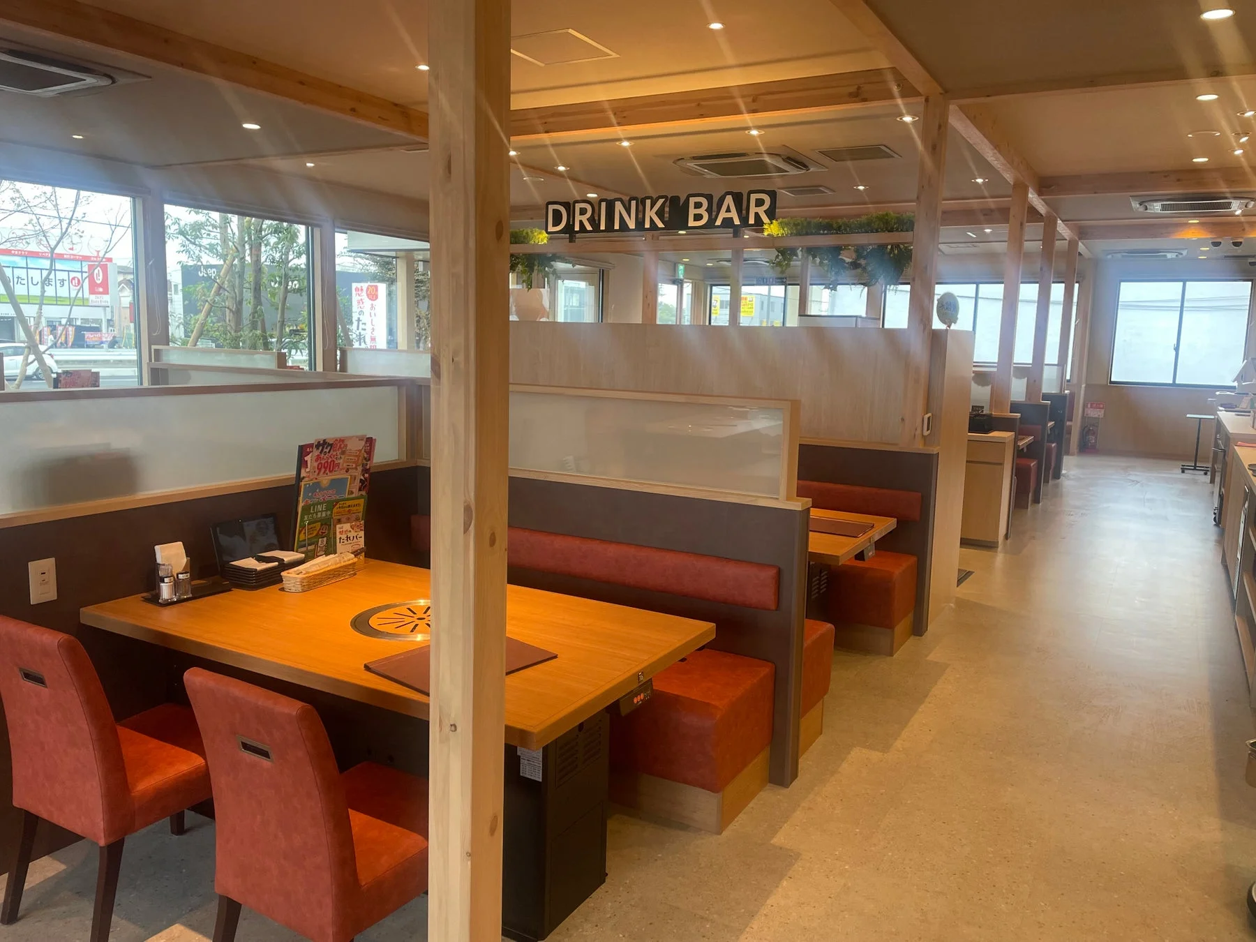 明るくモダンな雰囲気の焼肉店のような飲食店内部です。オレンジ色のクッションのブース席と木製テーブルが並び、「DRINK BAR」の表示が見えます。窓からは外の景色が望めます。