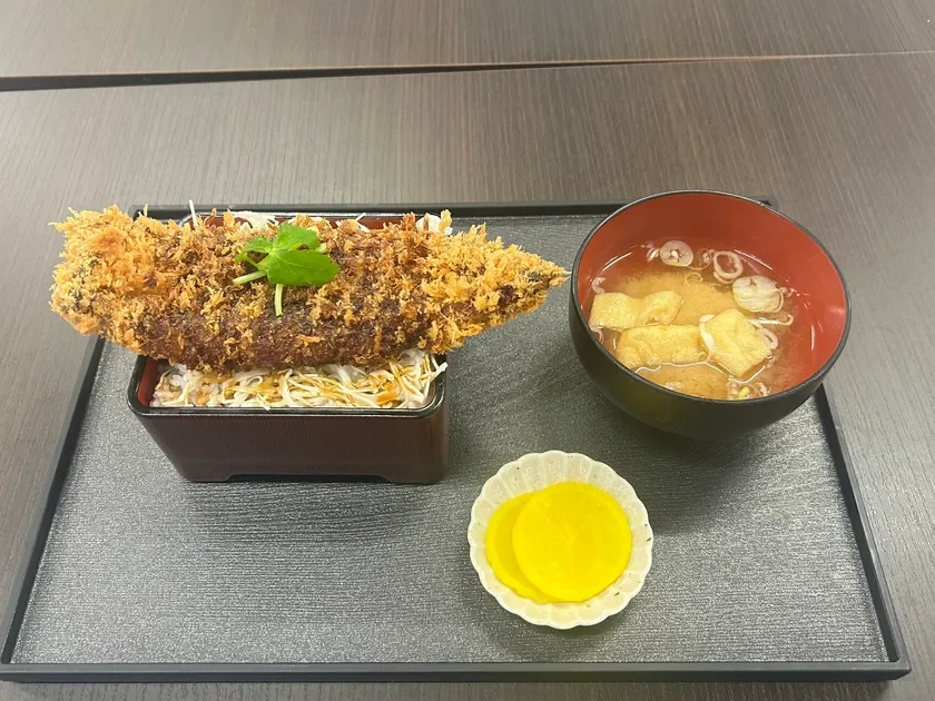 黒いお盆に載せられた和食の定食で、メインは大きな揚げ魚がご飯と千切りキャベツの上に盛り付けられています。他に味噌汁と黄色い漬物が添えられており、食欲をそそる一品です。