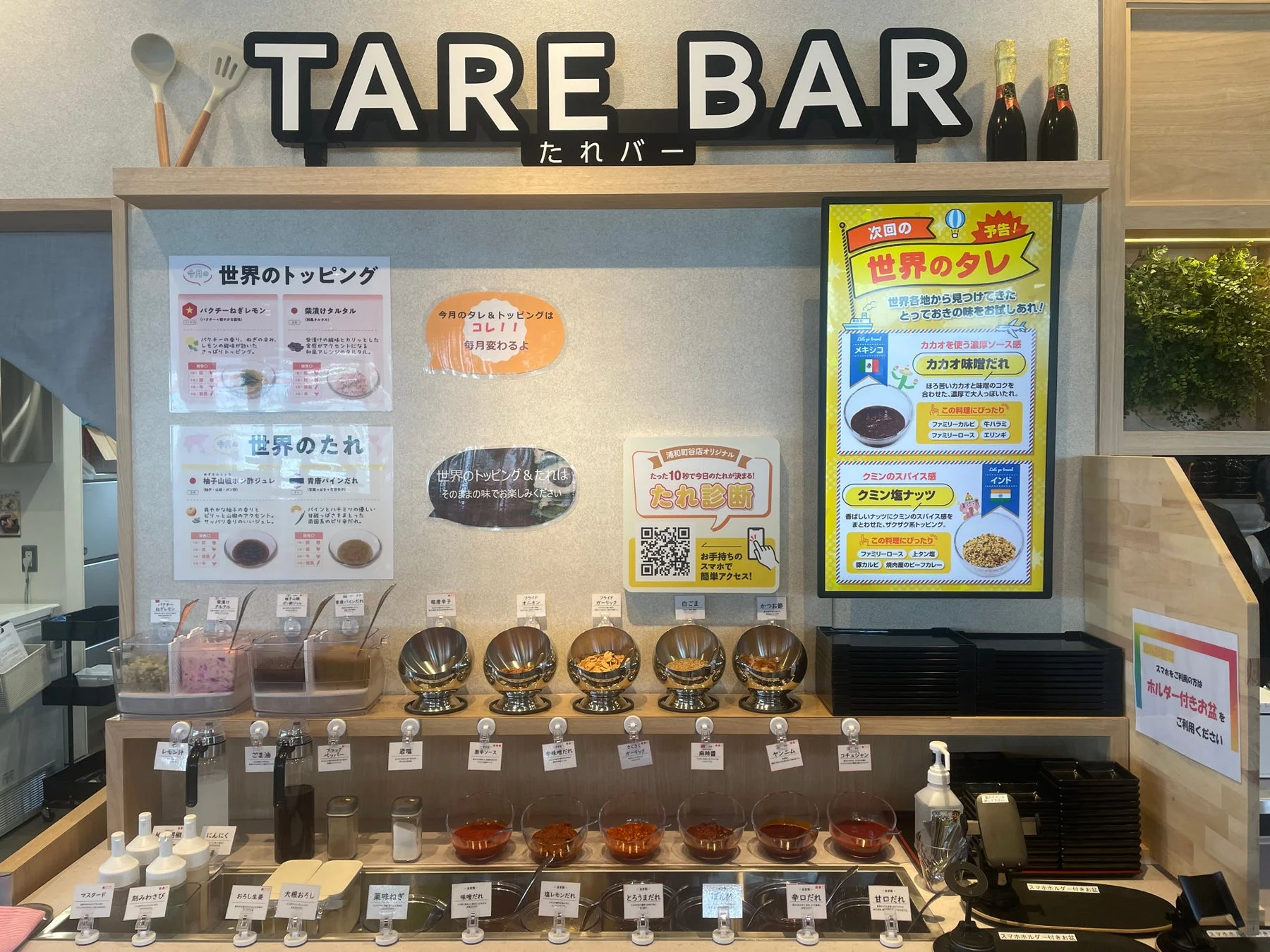 「TARE BAR」のセルフサービスコーナーで、世界のタレや今月のトッピングとして、カカオ味噌だれやクミン塩ナッツなど多種多様な調味料が並ぶ。スマホで最適なタレを選べる「たれ診断」も提供されている。