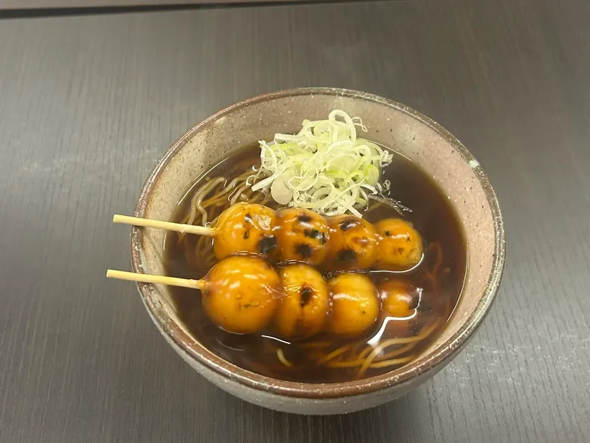温かい出汁に浸かった蕎麦の上に、香ばしく焼かれた団子が串に刺さって盛られています。薬味として刻みネギが添えられており、食欲をそそる和食の一品です。