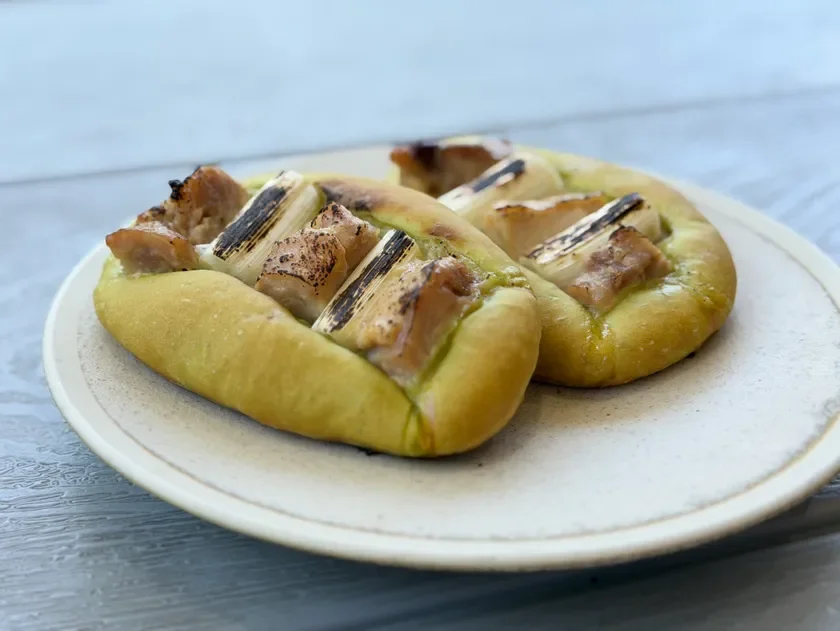 白い皿に盛り付けられた、焼き鳥とネギがトッピングされた惣菜パンが2つ。香ばしく焼き上げられた具材が特徴的な、食欲をそそる一品です。