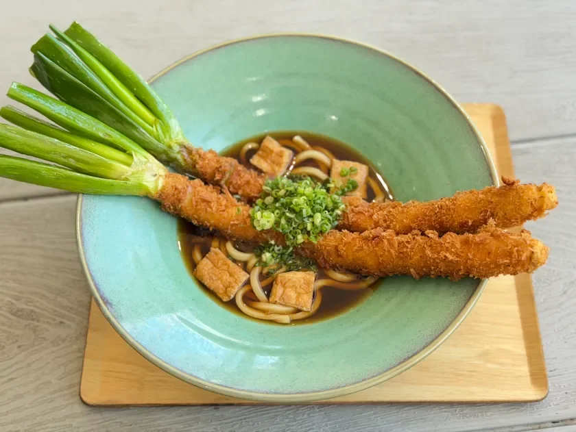薄緑色の器に入ったうどんです。だし汁に浸されたうどんの上に、大きなネギフライが2本と油揚げ、刻みネギがトッピングされています。木製のトレイに置かれ、温かそうな和食の様子が伝わります。