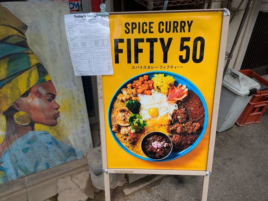 店の前に設置された「SPICE CURRY FIFTY 50」の黄色い看板がメインの画像です。看板には彩り豊かなスパイスカレーの大きな写真と「Today's Menu」と書かれたメニュー表が貼られています。看板の左隣には、ターバンを巻いた女性の絵画が立てかけられています。