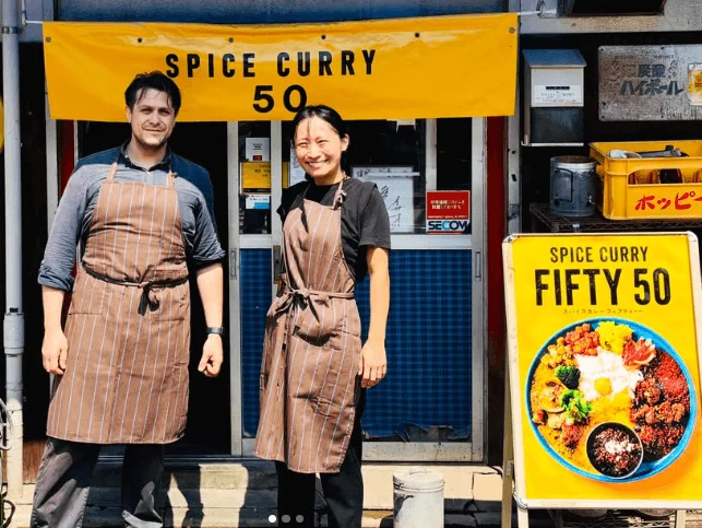 スパイスカレー店の前に立つ男女の店員。黄色い看板には「SPICE CURRY FIFTY 50」と書かれ、食欲をそそる彩り豊かなカレーの皿が描かれている。活気ある飲食店の日常風景を捉えた一枚。