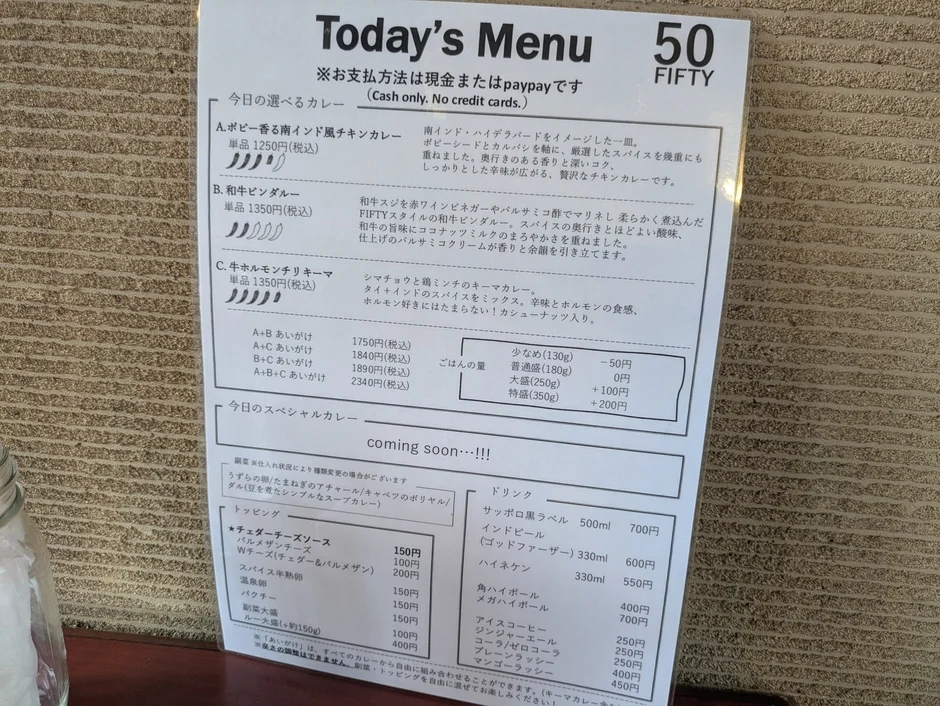 「FIFTY」の今日のメニューが写っています。南インド風チキンカレー、和牛ビンダルー、牛ホルモンチリキーマの3種類のカレーと、あいがけ、ご飯の量、トッピング、ドリンクが記載されています。支払いは現金かPayPayのみです。